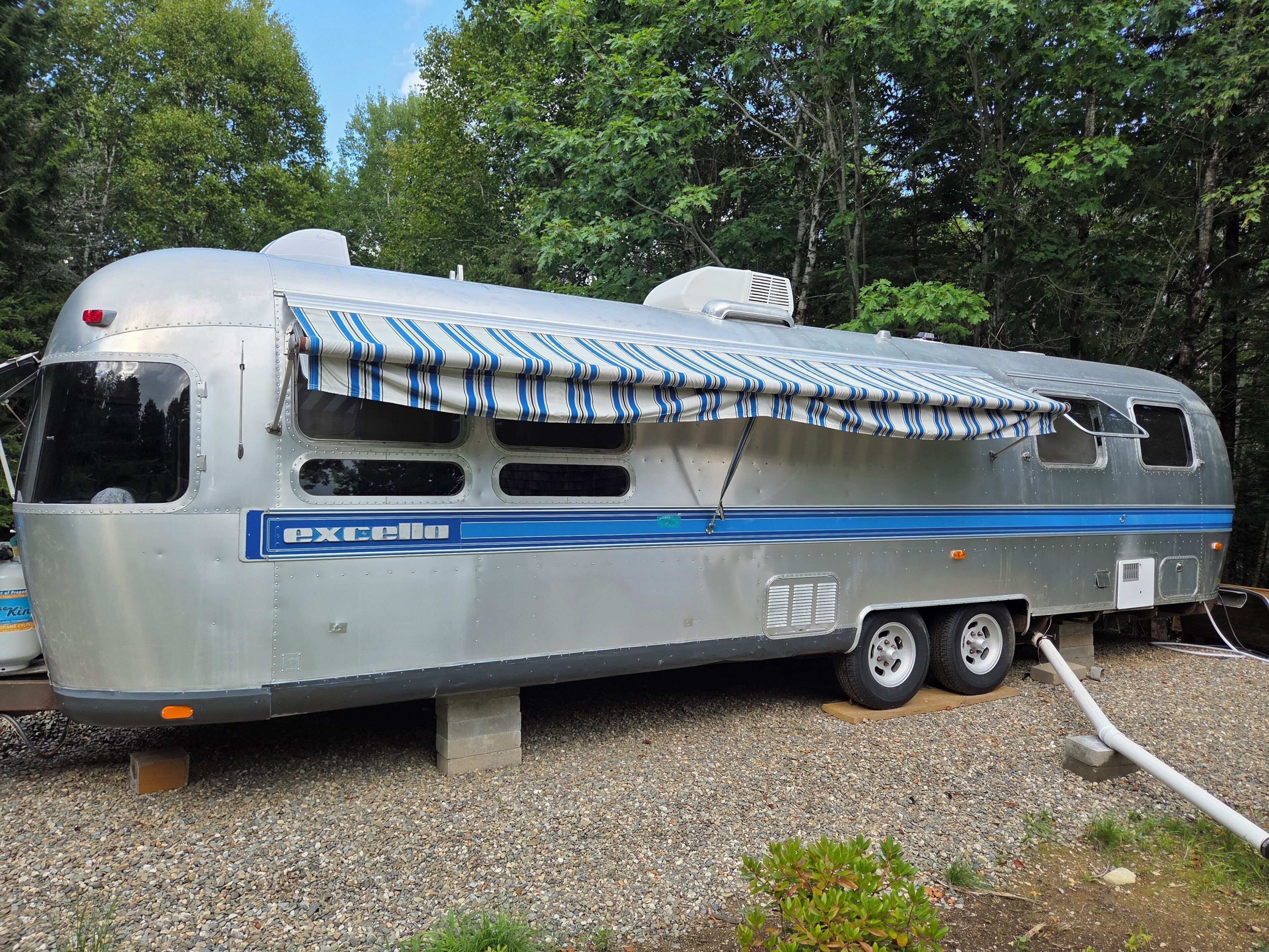Een zilverkleurige camper met een gestreept luifel, geparkeerd op grind met bomen op de achtergrond.