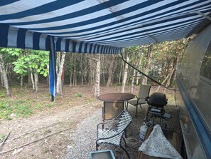 A campsite with a striped canopy, a round table, a grill, and a chair. - Vintage Airstream Excella in a Private Setting - Avail. May 15 through Oct. 31 (Penobscot)