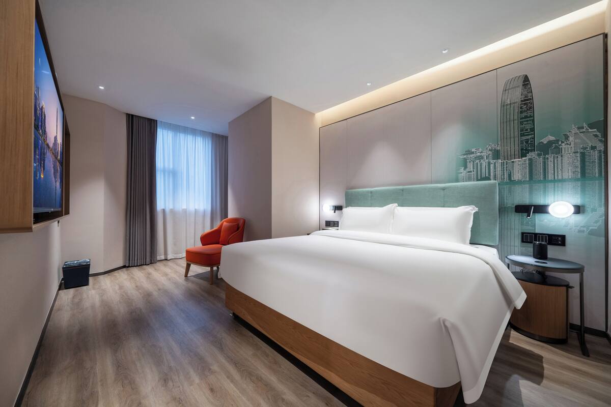 A modern hotel room with a large bed, a red chair, a cityscape wall art, and a nightstand with a lamp.