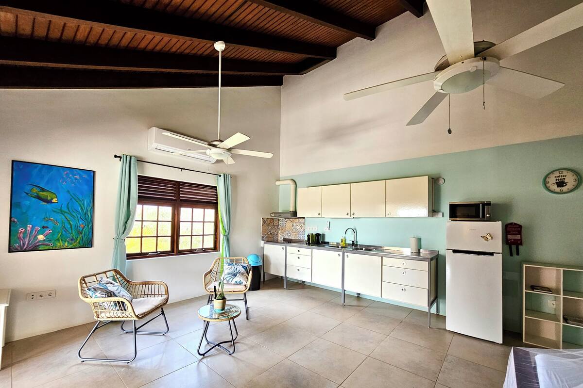 A spacious kitchen with white cabinets, a microwave, and a refrigerator. There are two ceiling fans, a window with curtains, and a wall-mounted clock.