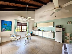 Una cocina amplia con armarios blancos, un microondas y un refrigerador. Hay dos ventiladores de techo, una ventana con cortinas y un reloj de pared.