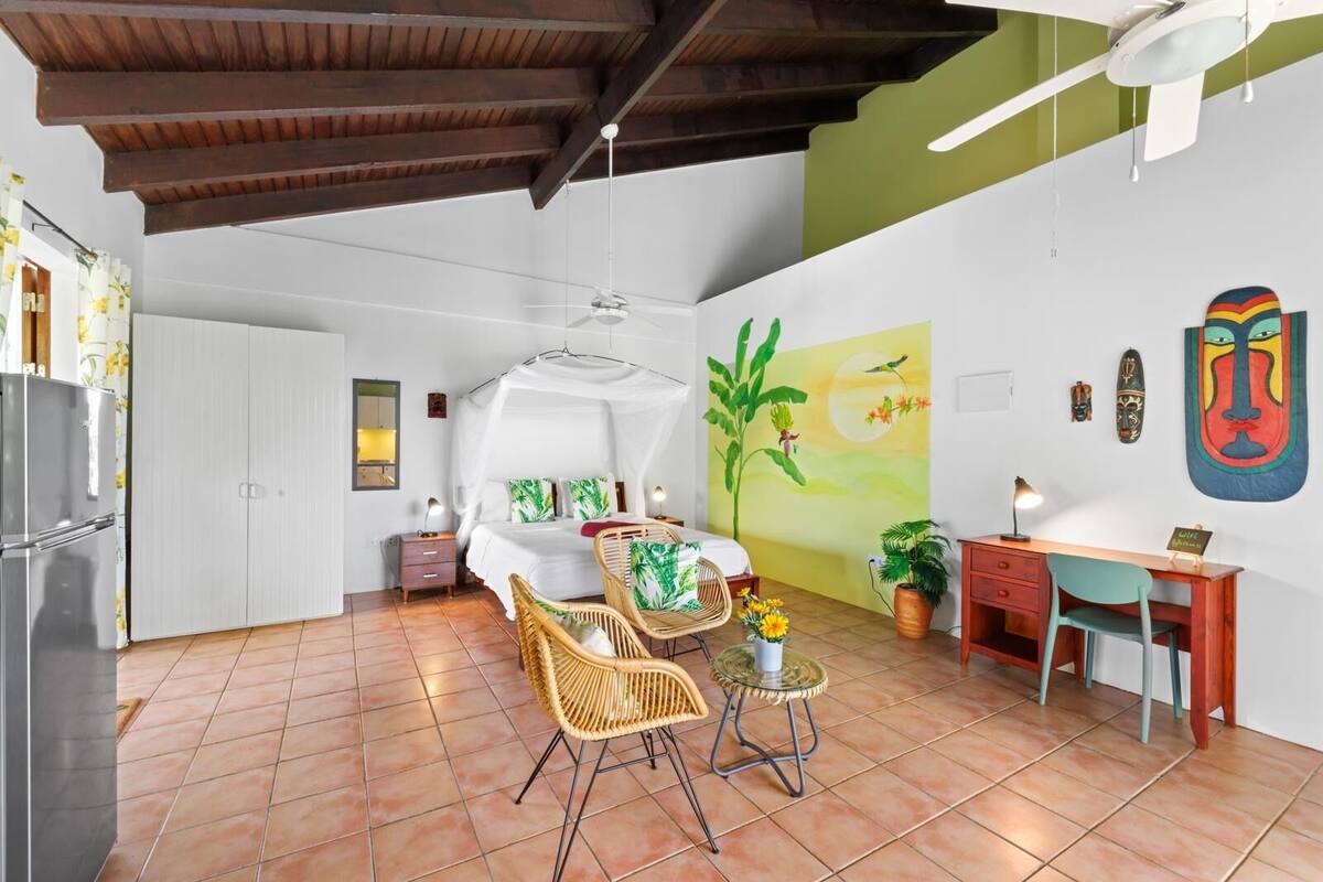 A room with a bed, a dining area with chairs, a table, a ceiling fan, and a wall with artwork.