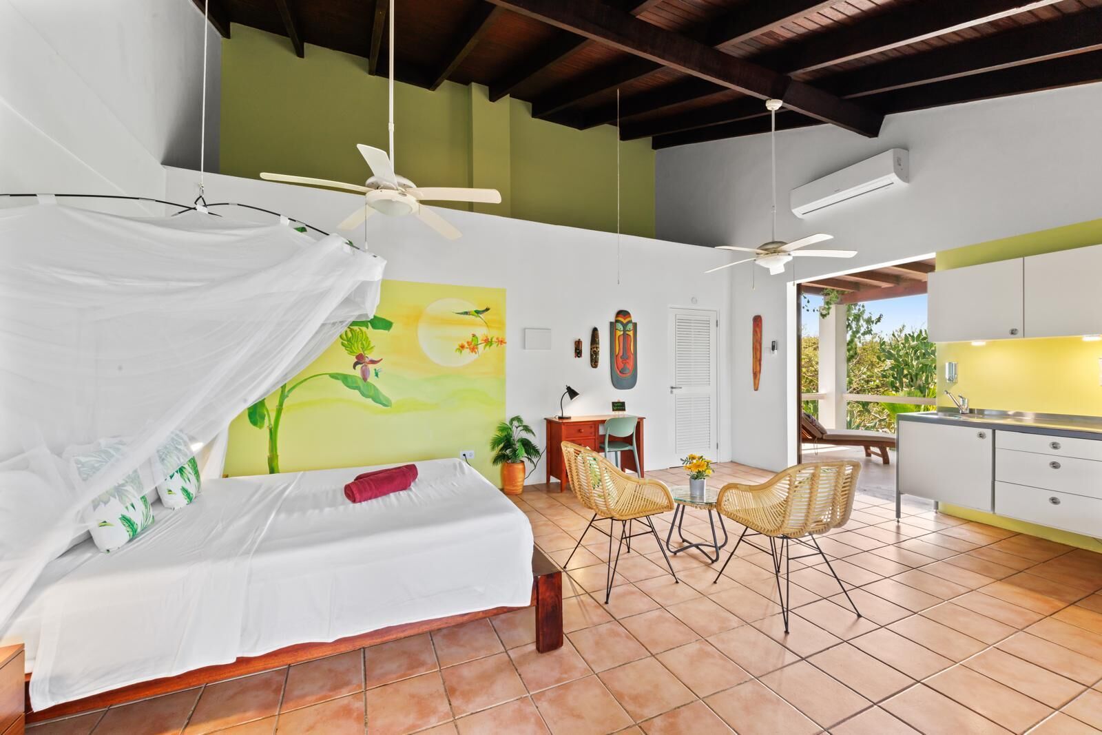 A bedroom with a bed, a desk, two chairs, a ceiling fan, and a view of the outside.
