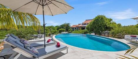 A swimming pool with lounge chairs and an umbrella, surrounded by greenery and a house in the background.