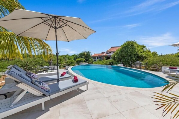 A swimming pool with lounge chairs and an umbrella, surrounded by greenery and a house in the background.
