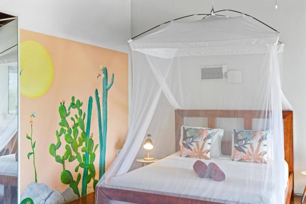 A room with a bed under a canopy, a cactus mural, and a nightstand with a lamp.