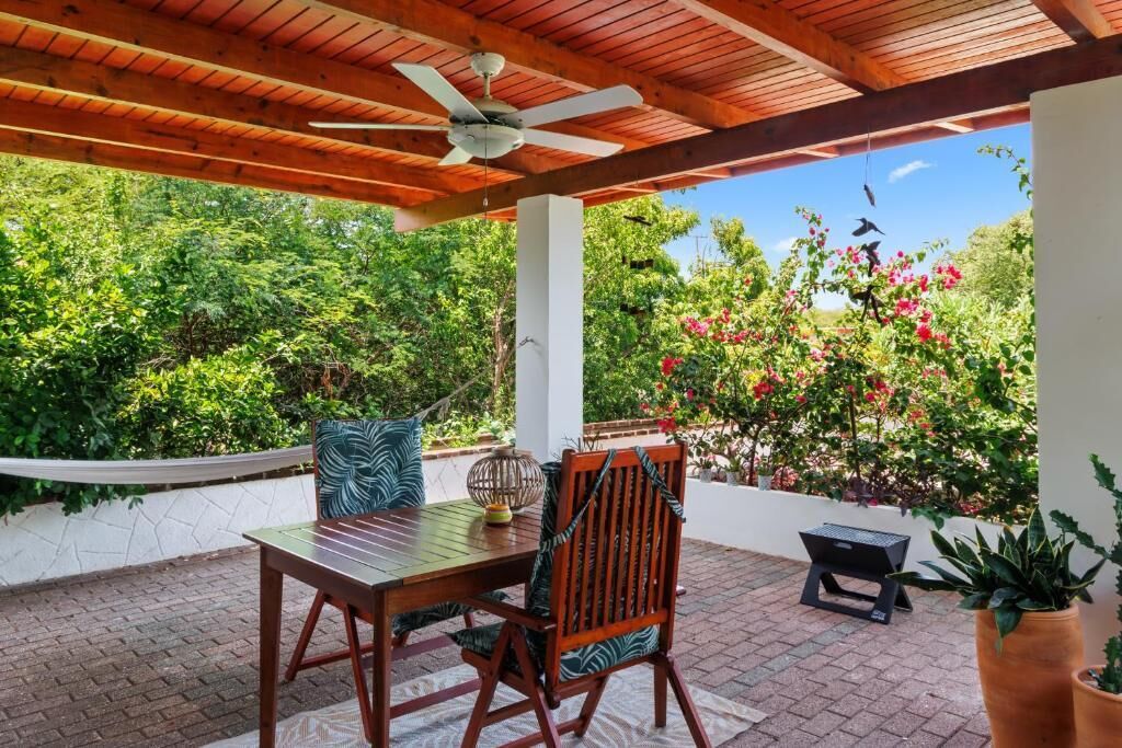 An outdoor patio with a wooden table and chairs, a ceiling fan, and a view of lush greenery and flowers.