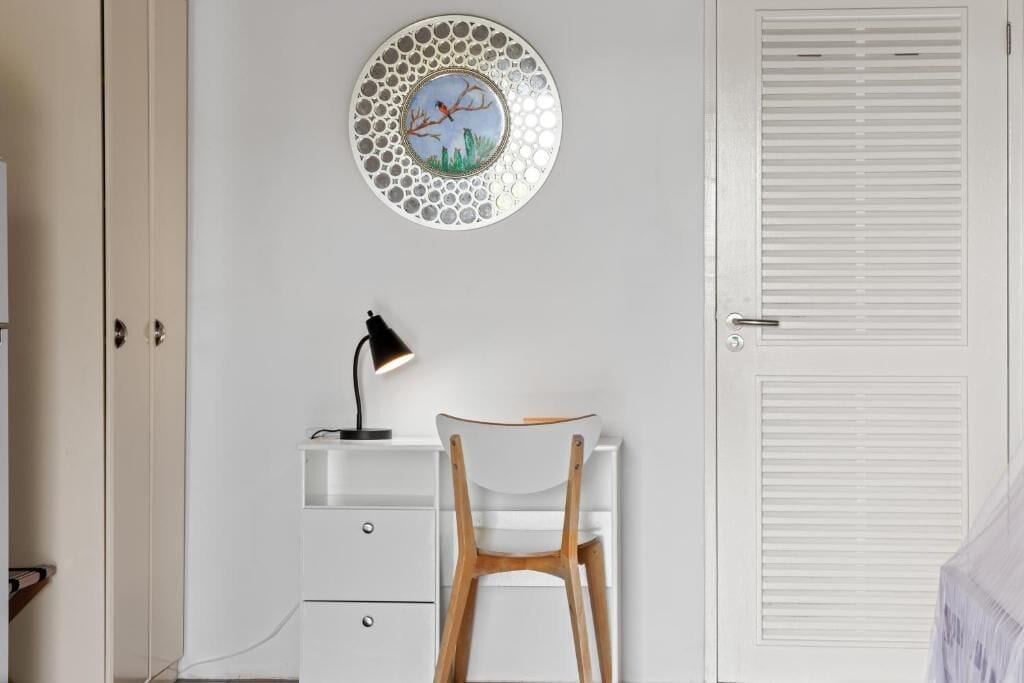 Una habitación con un escritorio blanco, una silla, una lámpara de pared y una puerta con paneles verticales.