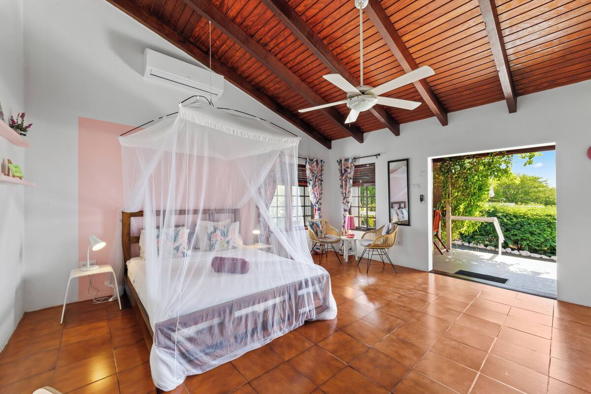 A bedroom with a bed under a canopy, a ceiling fan, a small table with a lamp, and a view of greenery outside.