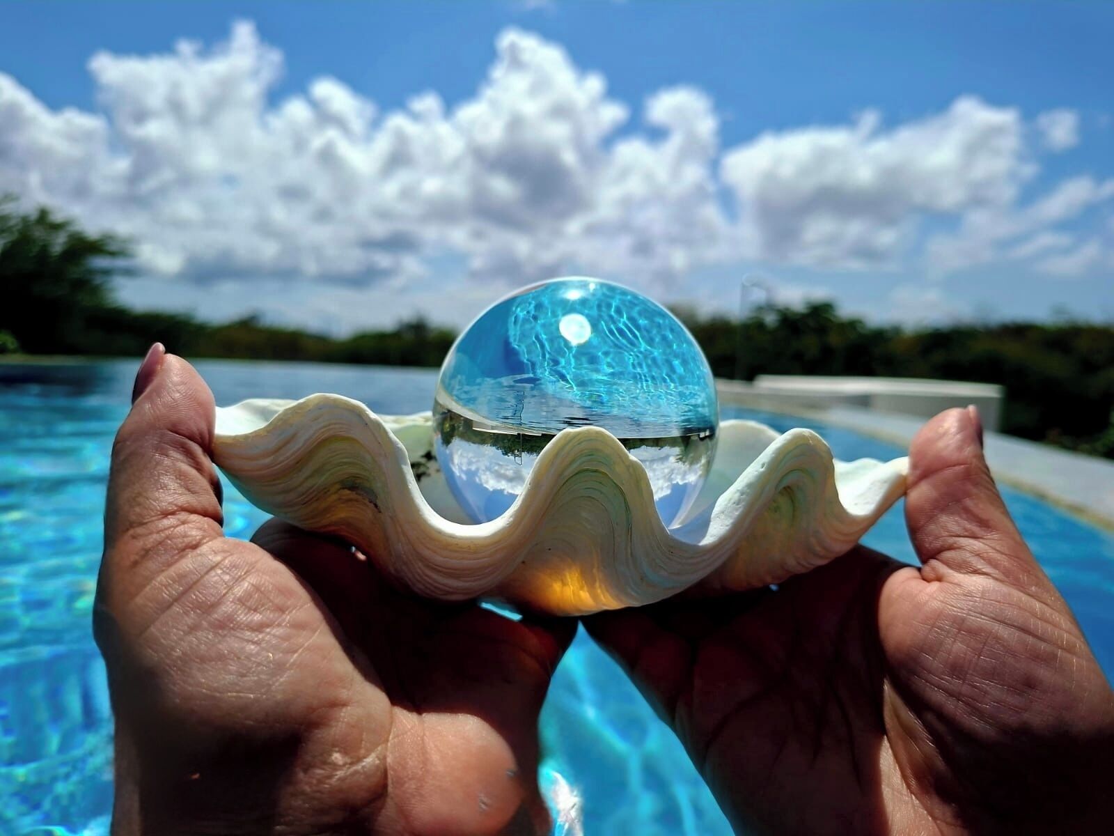 Une main tenant un bol en verre transparent à la surface réfléchissante, au-dessus d’une étendue d’eau.