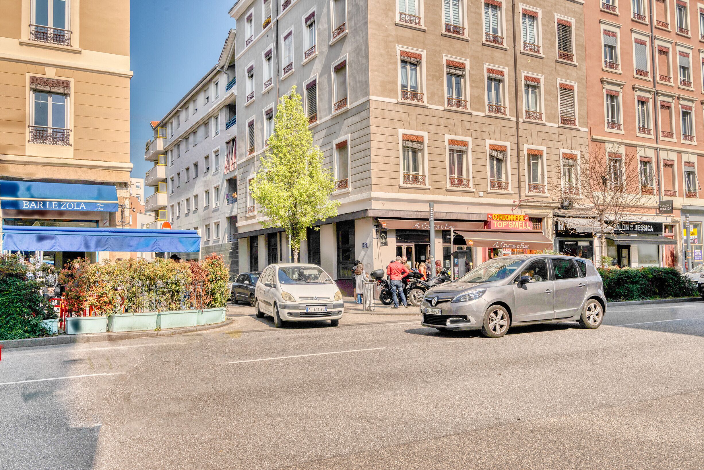 Une vue d’une rue, avec des voitures garées le long du trottoir, un bâtiment portant une enseigne indiquant « BAR LE ZOLA », et une autre enseigne indiquant « A. C. CORONERIE TOP’S SMELLIES ».
