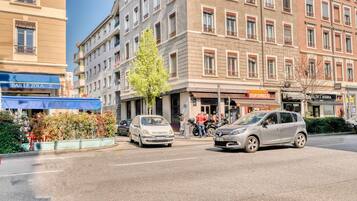 Une vue d’une rue, avec des voitures garées le long du trottoir, un bâtiment portant une enseigne indiquant « BAR LE ZOLA », et une autre enseigne indiquant « A. C. CORONERIE TOP’S SMELLIES ».