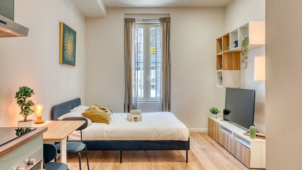 Une chambre à coucher avec un lit, une table de chevet, une lampe, une télévision et une fenêtre avec des rideaux.