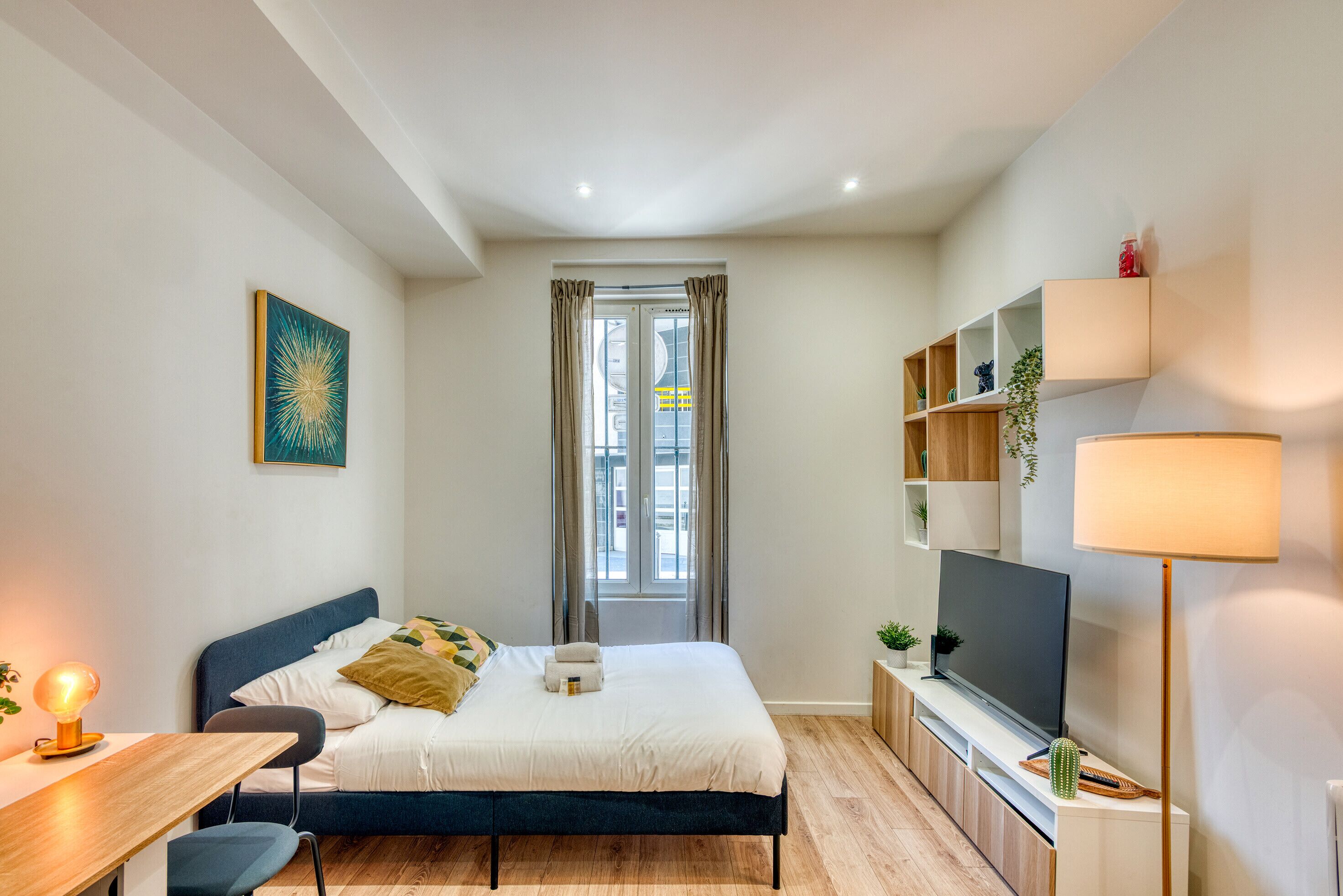 Ein Schlafzimmer mit Bett, Schreibtisch, Fernseher, Regalen und einem Fenster.