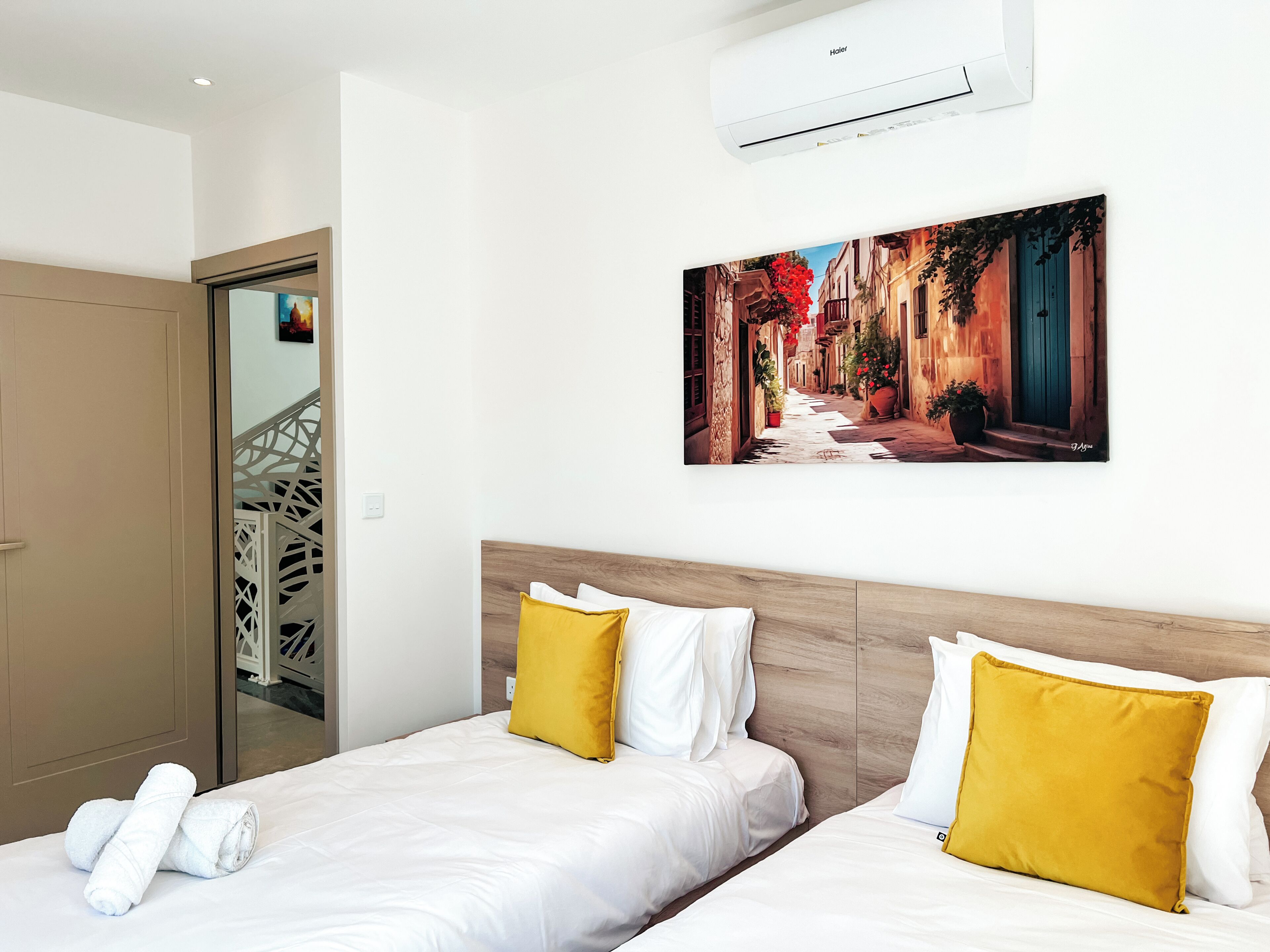 A hotel room with two beds, white bedding, yellow pillows, a painting of a street scene, and an air conditioning unit.