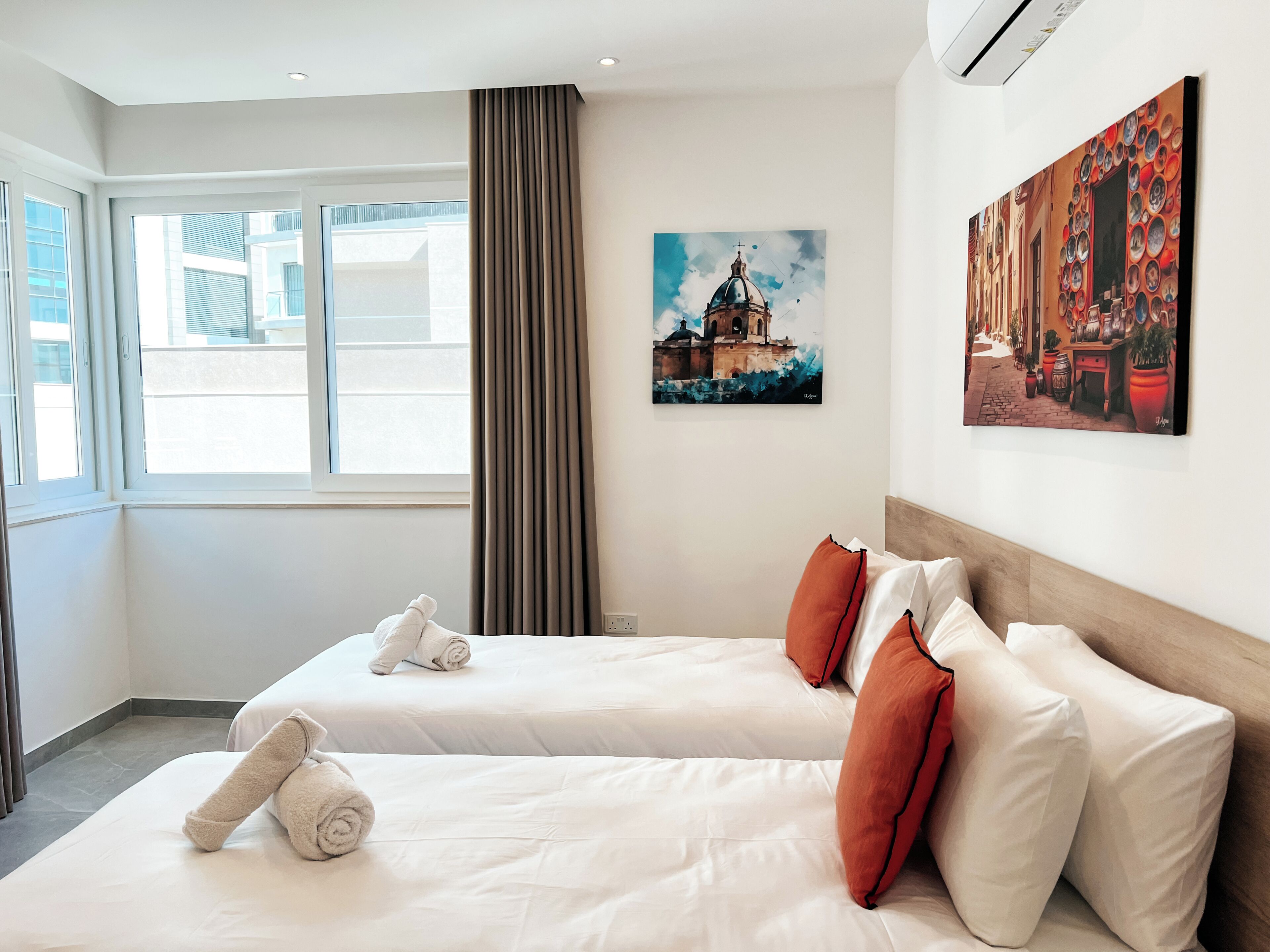 Una habitació d'hotel amb dos llits, una finestra gran, un quadre d'un edifici i un aparell d'aire condicionat.