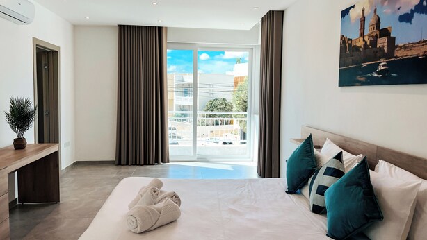 A hotel room with a bed, a desk, a potted plant, a painting, and a view of the city.
