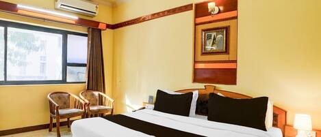 Habitación de hotel con una cama grande, dos sillas, una mesita de noche, aire acondicionado, una ventana con cortinas y un cuadro colgado en la pared.