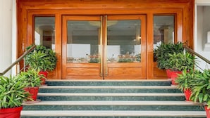 Une porte à deux battants en bois, ornée de panneaux de verre, flanquée de jardinières rouges où poussent des plantes vertes, et donnant sur un escalier aux marches en marbre.