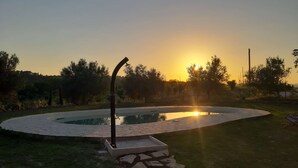 Uma piscina circular com borda de pedra, cercada por grama e árvores, com o sol se pondo ao fundo.