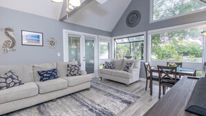 A modern living room with a gray sofa, a dining table, and a large window with a view of greenery. - Your Shore Drive Escape Awaits at Arcadian Dunes 27-609 (Myrtle Beach)