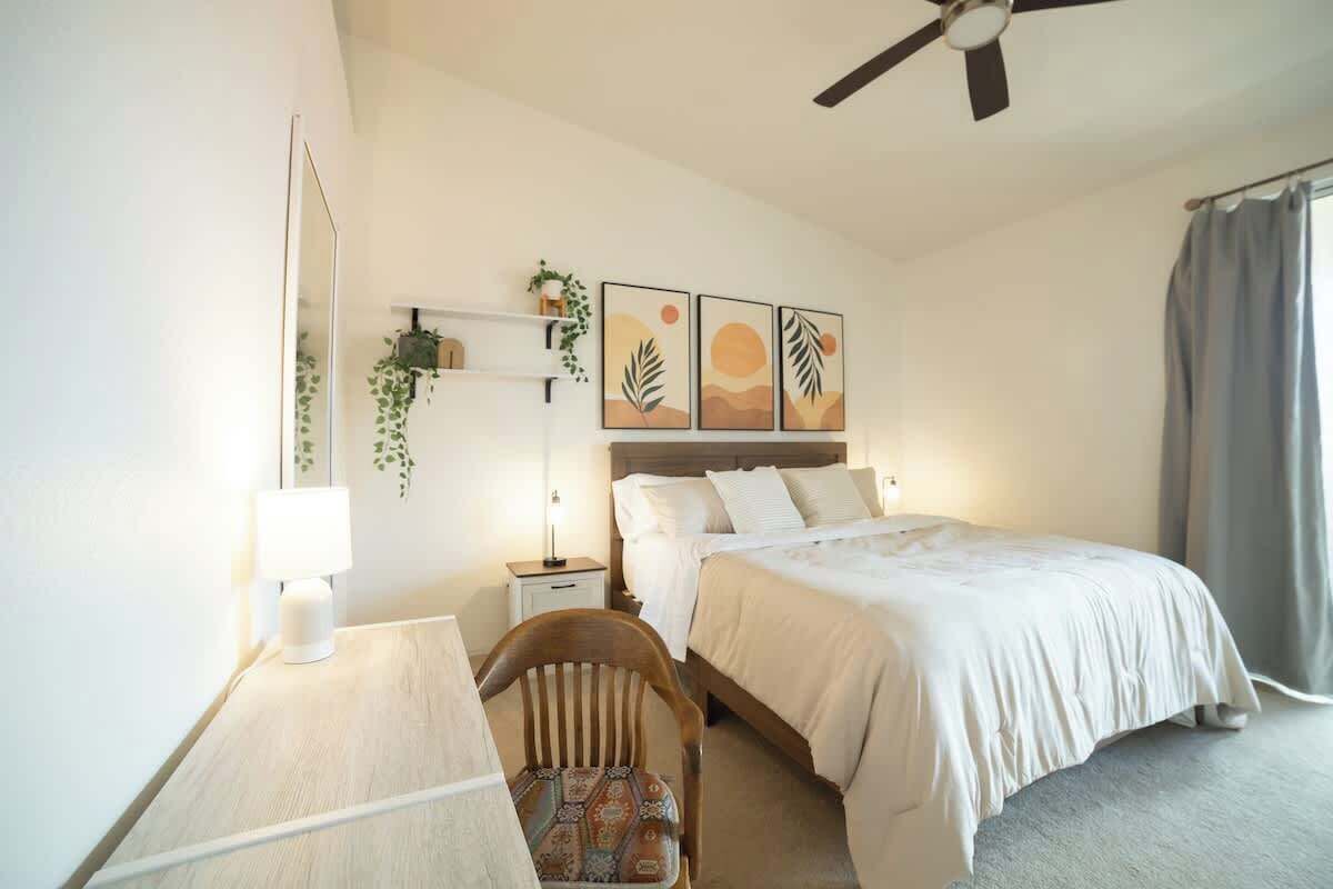 A bedroom with a bed, a desk with a chair, a bedside table, a lamp, a ceiling fan, and wall art.