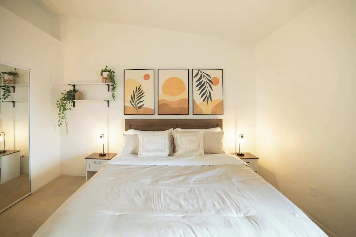 A bedroom with a bed, bedside tables, a nightstand, a mirror, and wall-mounted shelves with plants.