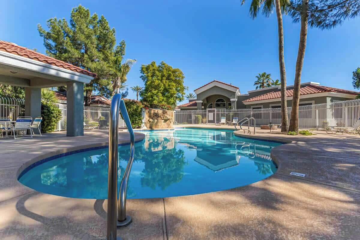A swimming pool with a curved edge, surrounded by a fence, and a covered seating area with chairs.