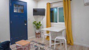 Une pièce avec une porte bleue, un bureau blanc avec deux chaises, une petite table, une plante en pot et un téléviseur fixé au mur.