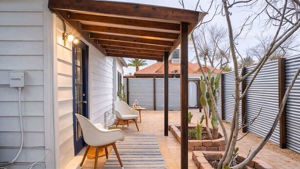 Une terrasse couverte avec deux chaises blanches, une pergola en bois et un jardin en briques où sont plantés de petits arbres.