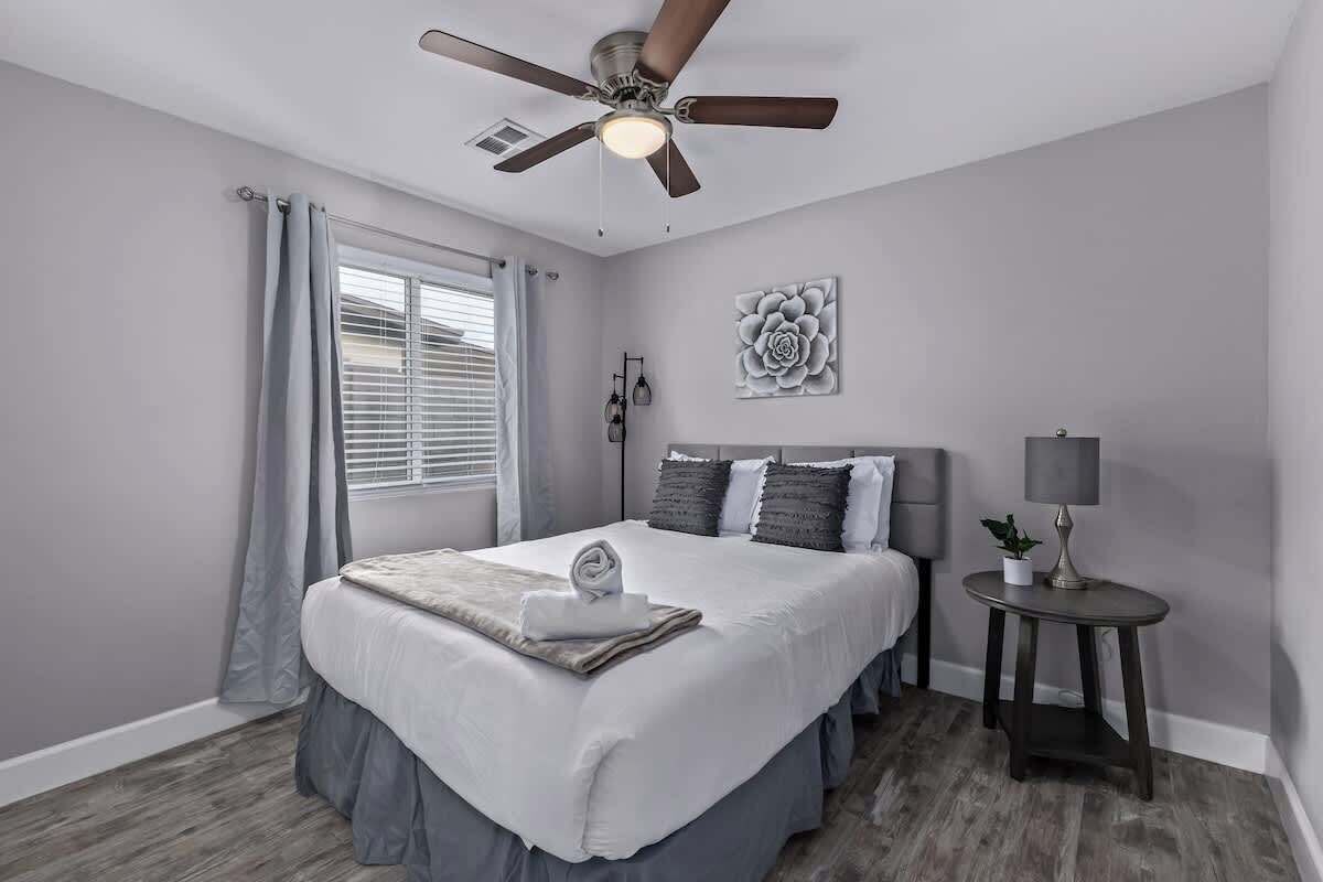 A bedroom with a bed, a ceiling fan, a bedside table with a lamp, and a window with curtains.