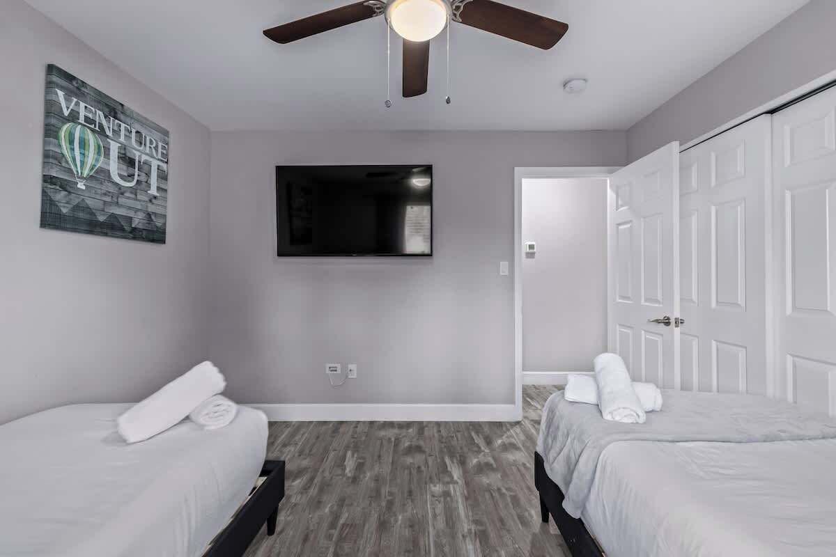 A bedroom with two beds, a ceiling fan, a mounted TV, and a door leading to another room.