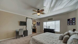 Ein Schlafzimmer mit Bett, Schreibtisch, Stuhl, Fernseher, Deckenventilator und einem Fenster mit Jalousien.