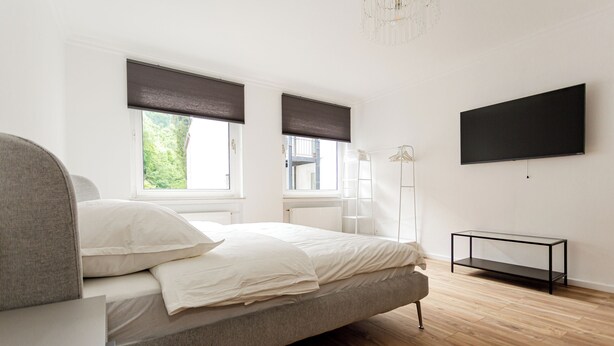 Un dormitorio con cama, dos ventanas con persianas, un televisor en la pared y suelo de madera.