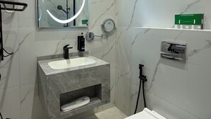 Ett badrum med en vit toalett, ett handfat med en bänkskiva i marmor, en spegel med en rund lampa och en duschhörna.
