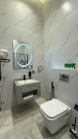 Een badkamer met een wit toilet, een wastafel met een marmeren blad, een spiegel met rondom verlichting, en een douchehoek.