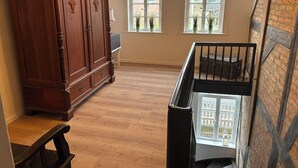 A room with a wooden floor, a large wooden cabinet, two windows with potted plants, and a staircase with a black railing. - Central and quiet: freshly renovated old apartment with charm (Helpsen)