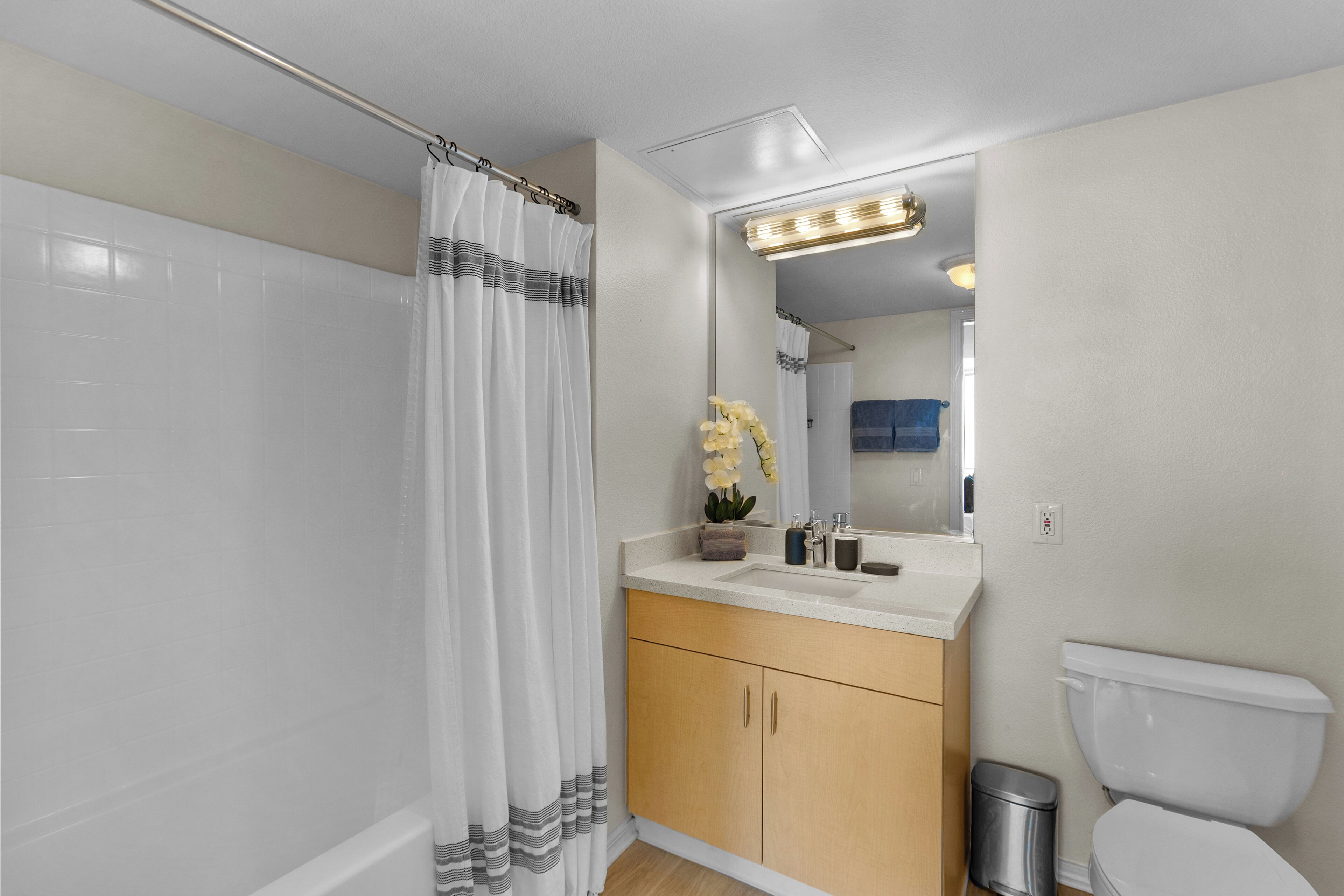 A bathroom with a shower curtain, a sink with a mirror above it, a toilet, and a trash can.