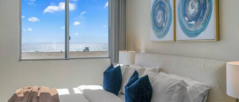 A hotel room with a bed, pillows, a view of the beach, and abstract art on the wall.