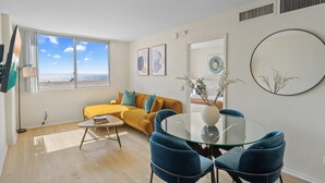 Uma sala de estar moderna com sofá, mesa de vidro e cadeiras, uma janela grande com vista e parede com obras de arte emolduradas.