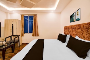 A hotel room with a bed, a desk, a chair, a wardrobe, a window with curtains, and artwork on the wall. - Deluxehotel Niraant (Prayagraj)