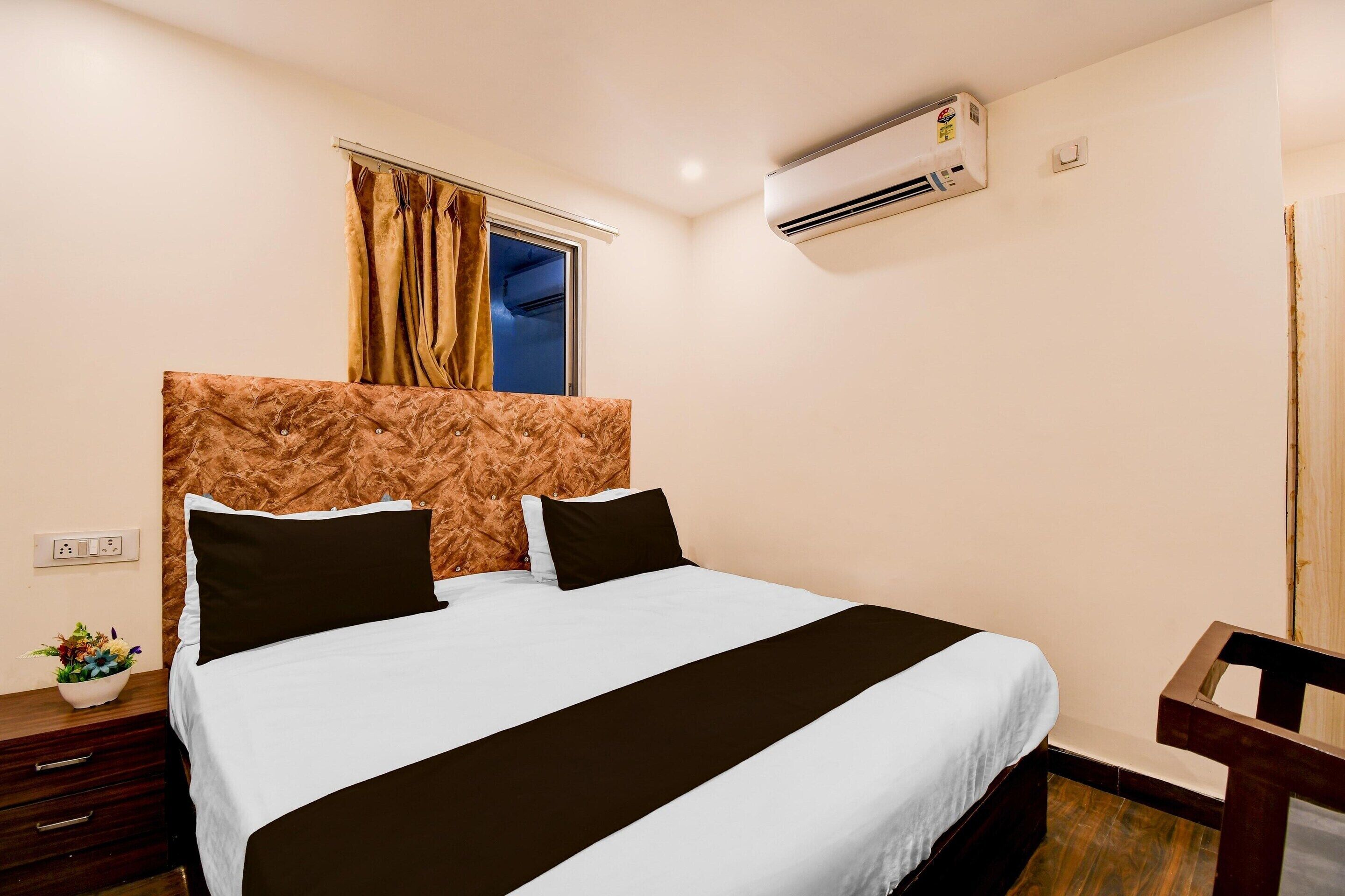 Quarto de hotel com cama, mesa de cabeceira, janela com cortinas, ar condicionado e uma cadeira de madeira.