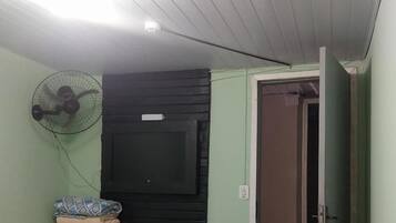 Una habitación con una cama, un mueble pequeño, un televisor montado en un panel negro, un ventilador de pie y una puerta.