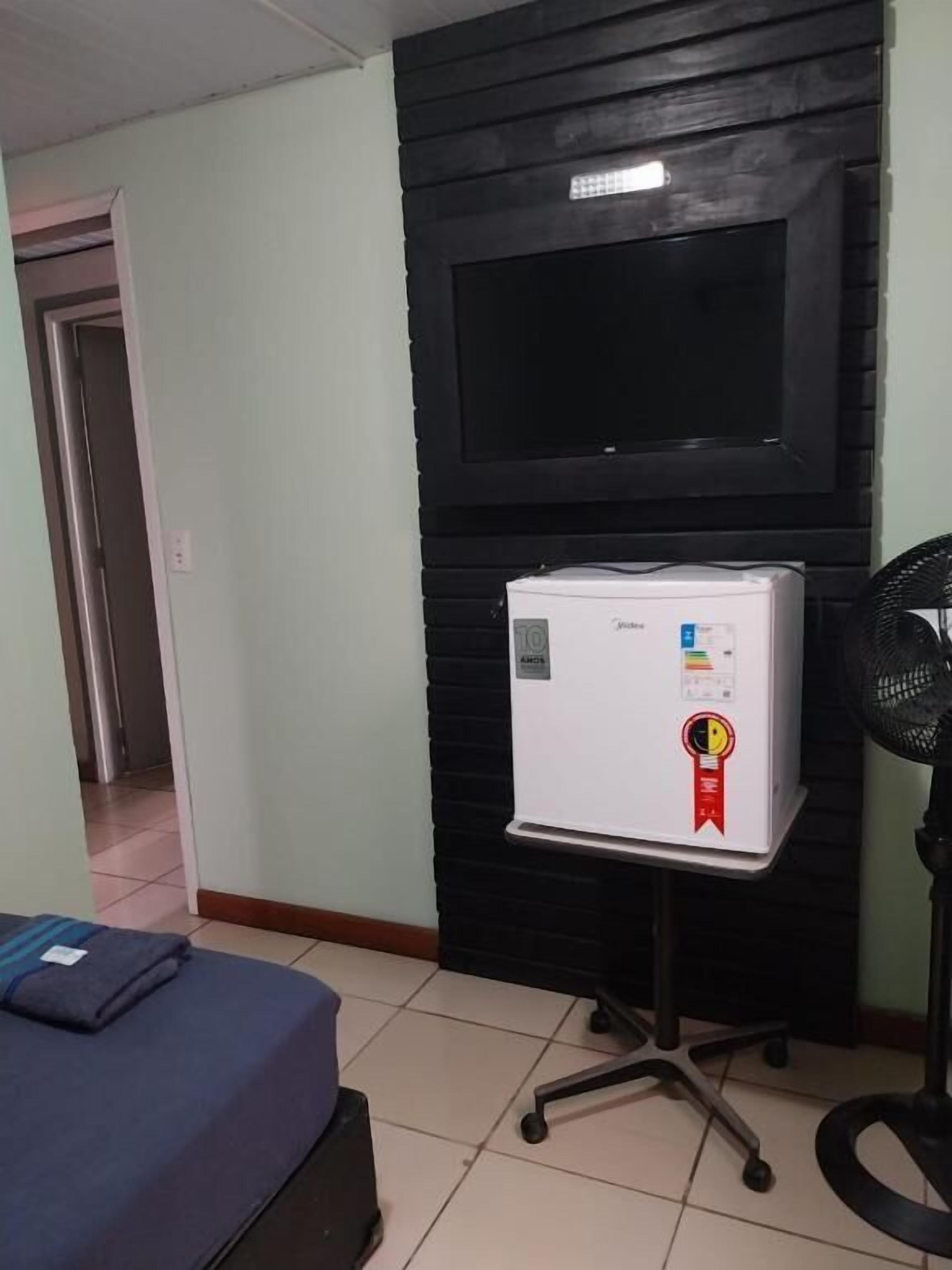 Una habitación con una cama, una pequeña mesa con refrigerador, un televisor montado en una pared negra y un ventilador de pie.