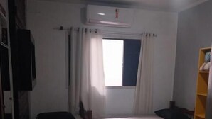 Ein kleiner, gut beleuchteter Raum mit zwei Betten, einem Vorhangfenster, einem an der Wand befestigten Fernseher und einem Regal mit Gegenständen.