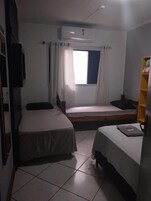 Quarto pequeno e bem iluminado, com duas camas, uma janela com cortinas, uma televisão na parede e uma estante com objetos.