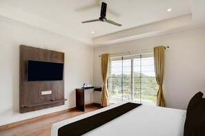 A hotel room with a bed, a TV mounted on the wall, a desk, and a window with curtains. - Deluxe Arasu Comforts (Hassan)