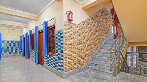 A hallway with a checkered floor, a staircase with a woven wall, and a fire extinguisher mounted on the wall. - Classic With Balcony Diamond Inn (Durgapur)