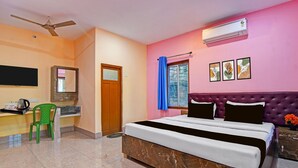 A hotel room with a bed, a desk with a chair, an air conditioner, a ceiling fan, a window with curtains, and artwork on the wall. - Classic With Balcony Diamond Inn (Durgapur)