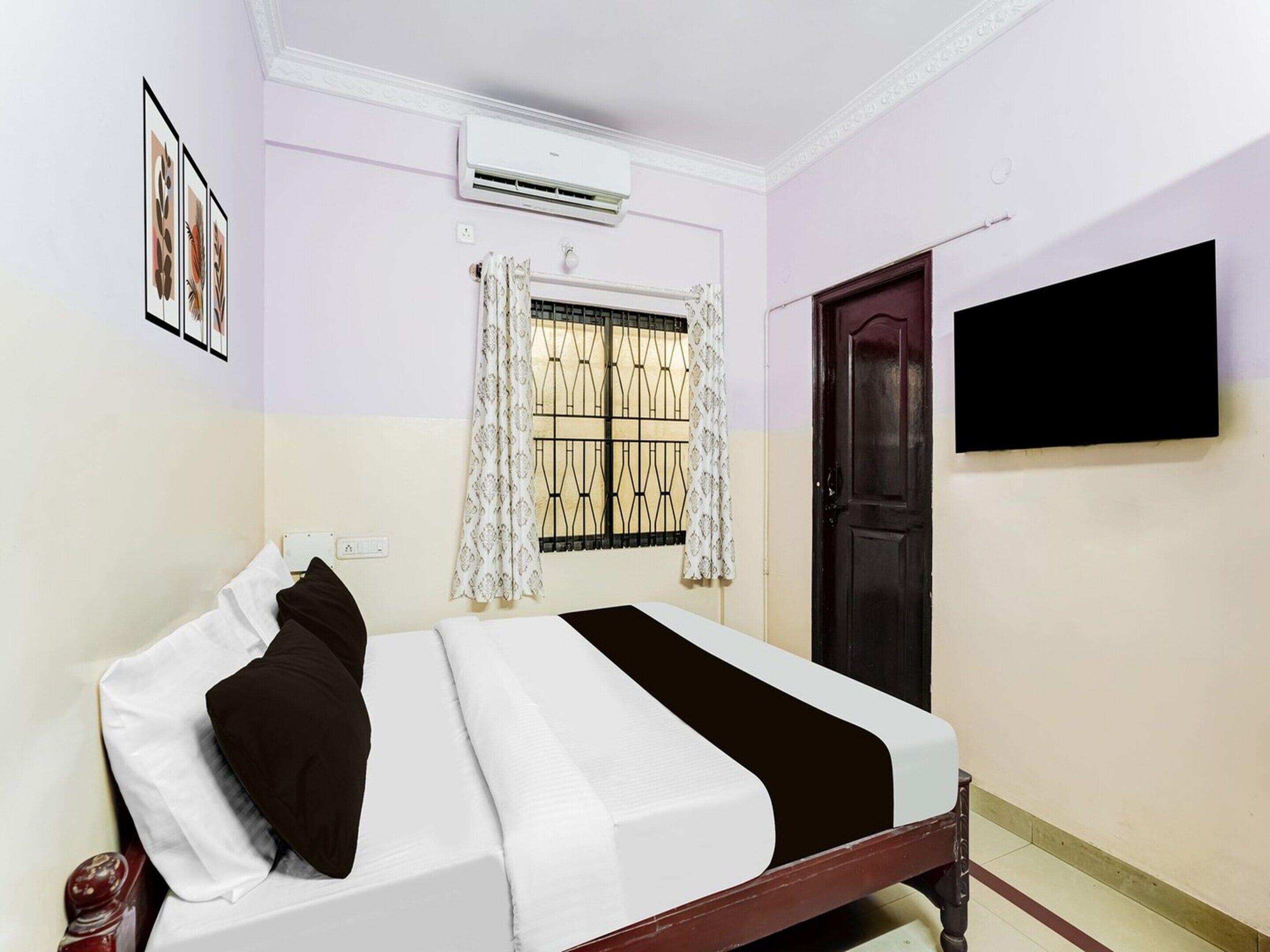 A bedroom with a bed, an air conditioner, a window with curtains, a door, and a wall-mounted TV.